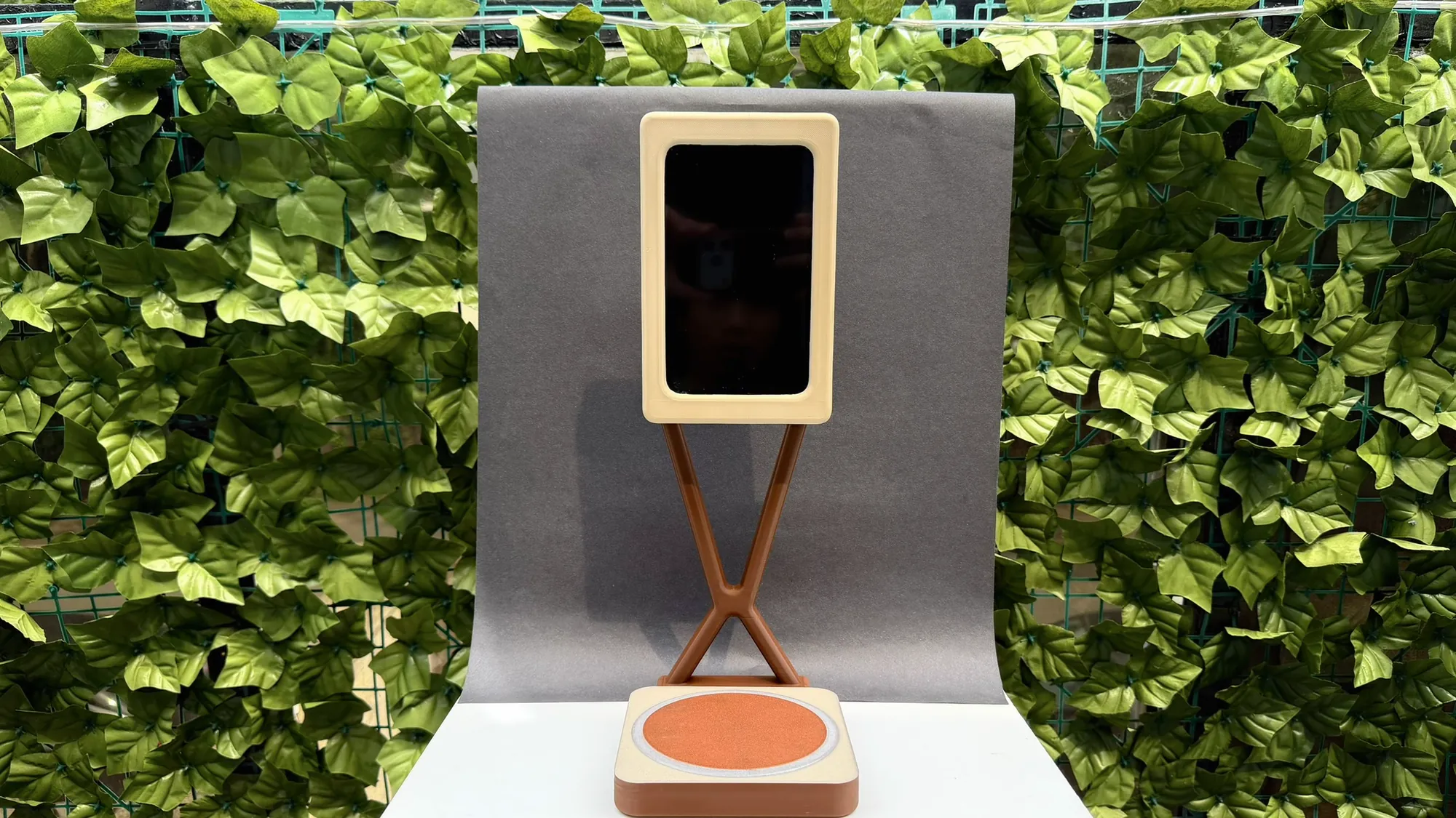 The finished BrewFlow prototype in front of a hedge: a cream screen frame on a brown X-shaped stand above a cream and brown base with a terracotta coaster