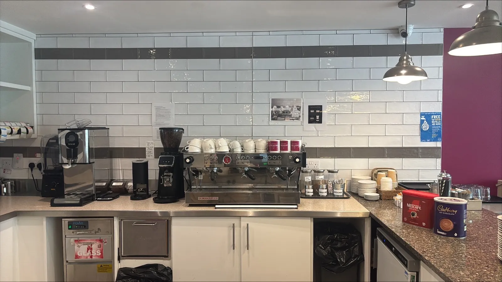 A small café counter with an espresso machine, grinder and white tiled wall