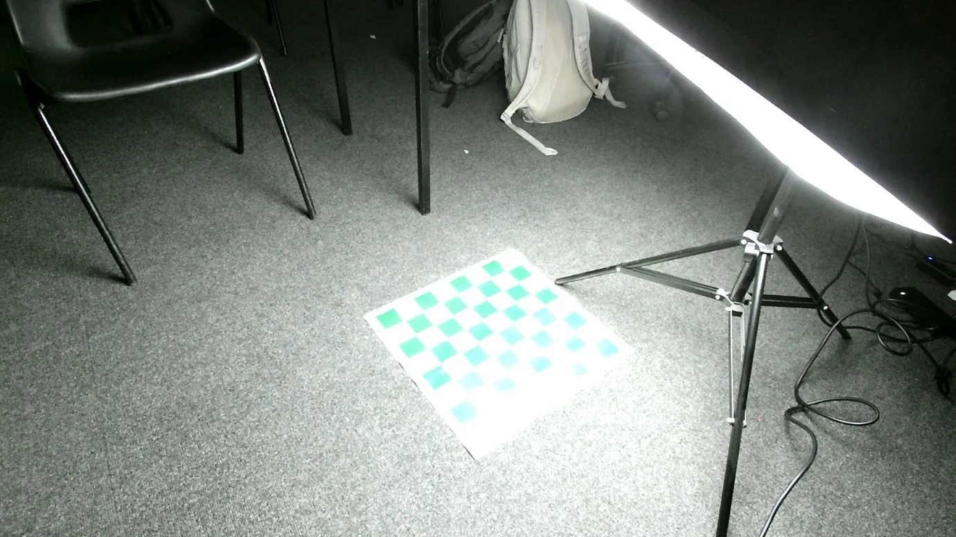 A printed green and white chessboard on a grey carpet, lit by a large studio softbox on a tripod, with a chair in the background
